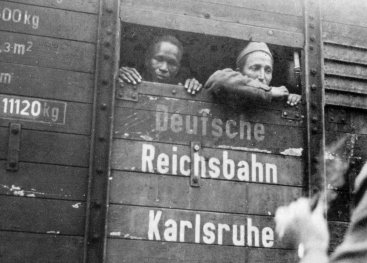 Two men  in a train wagon looking out of a small gap. On the wagon it says: 'Deutsche Reichsbahn'.